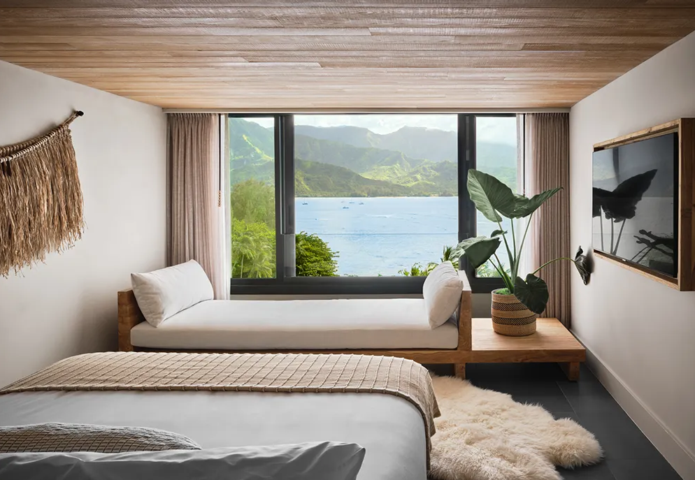 Cozy bedroom with a view of the water and mountains through a large window. Includes a bed, couch, and decor.