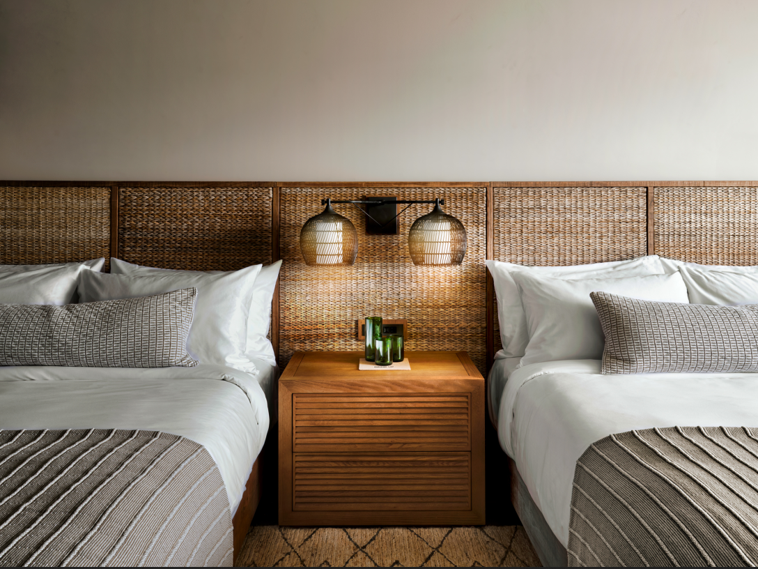 Twin beds with woven headboard, bedside table, and wall sconces in a hotel room.