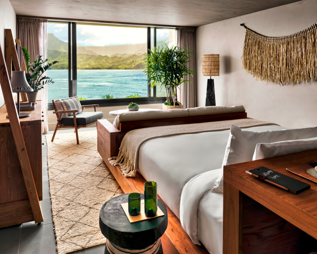 Hotel room with bed, desk, and ocean view through a large window. Neutral tones, natural materials, and a plant.