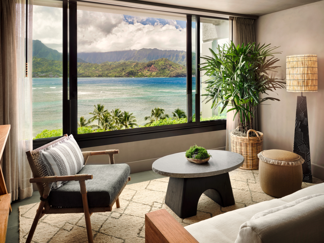 Living room with large window showcasing ocean view and mountain range. Furniture includes armchairs, coffee table, and lamp.