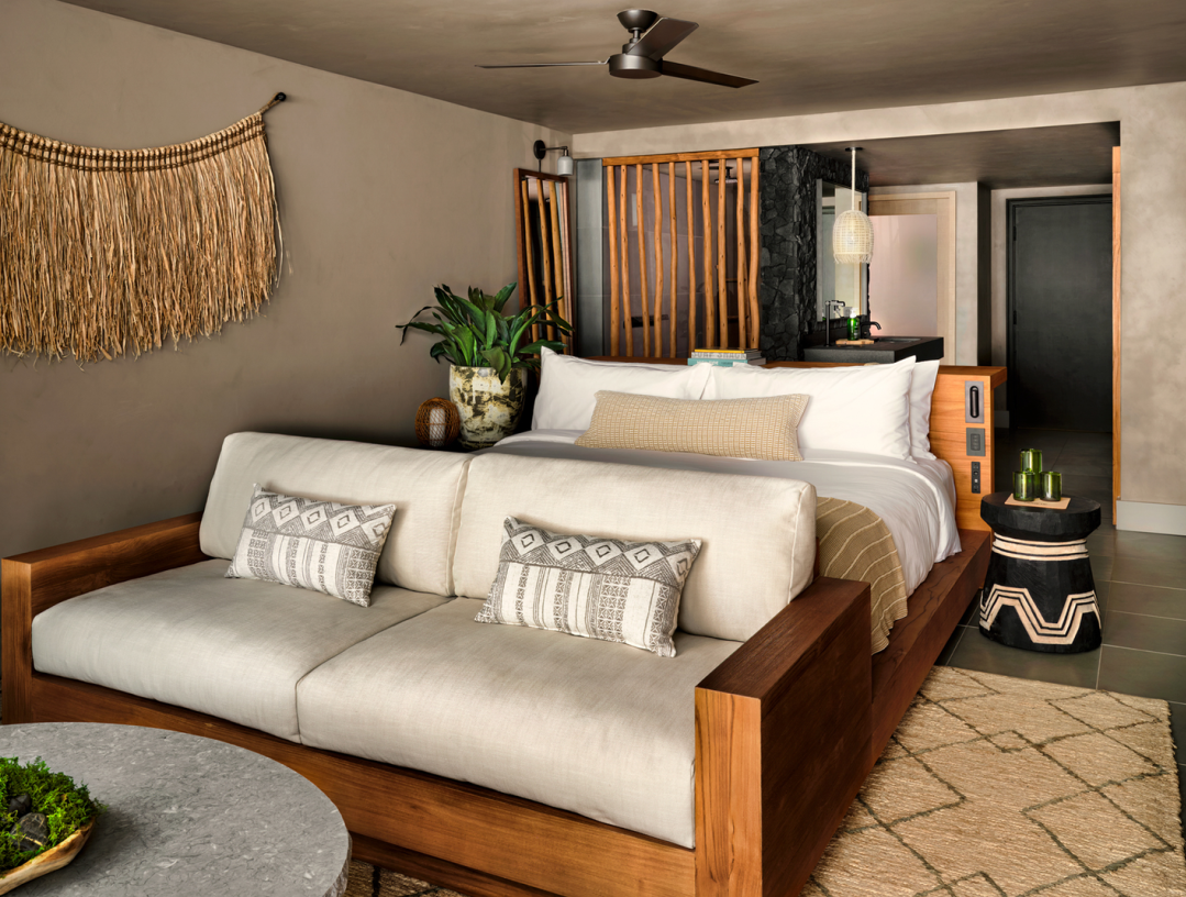 A modern hotel room with a bed, sofa, and woven wall hanging in earthy tones.