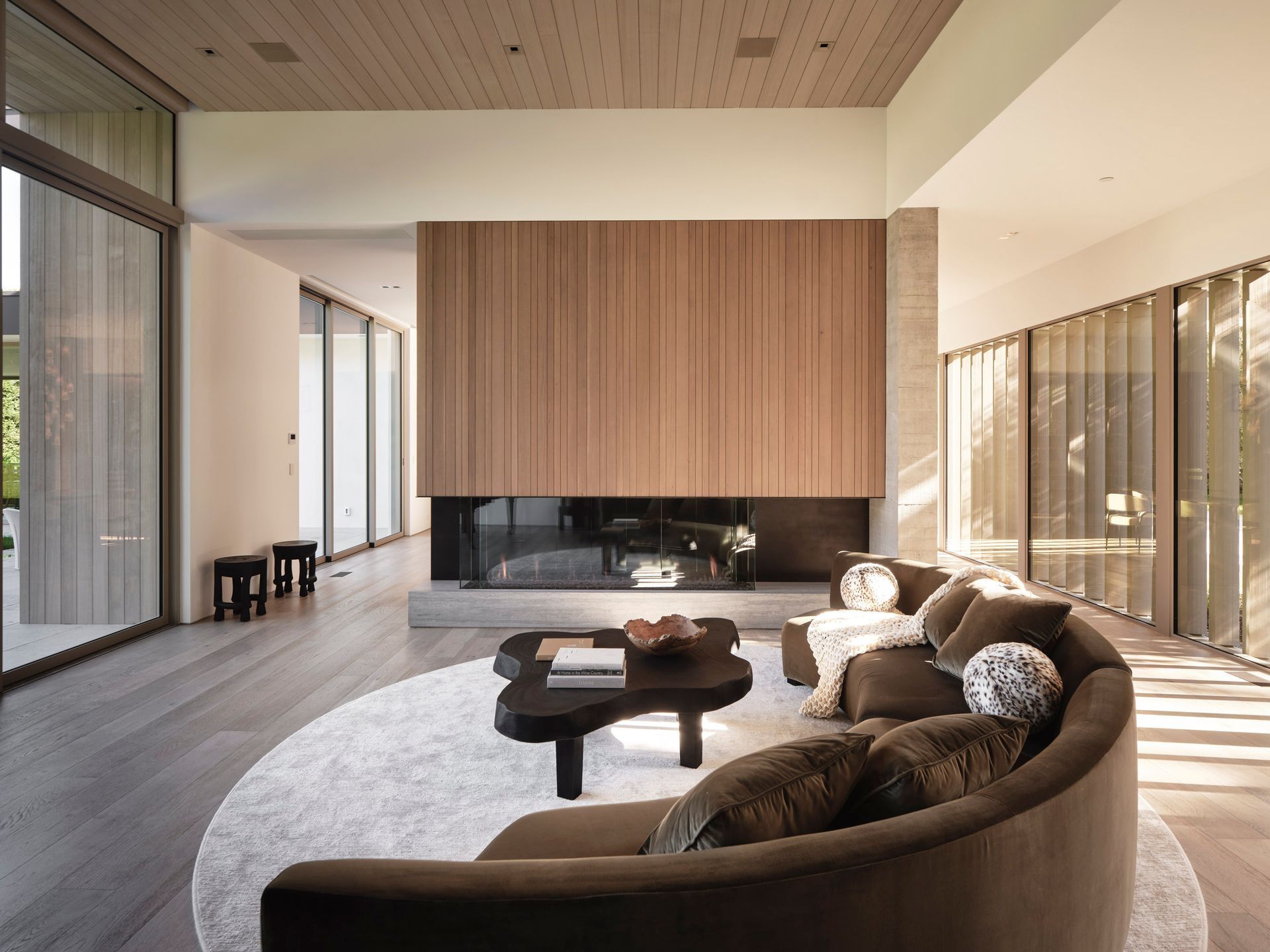 Modern living room with a brown curved sofa on a circular rug, a wood-paneled fireplace, and floor-to-ceiling windows.