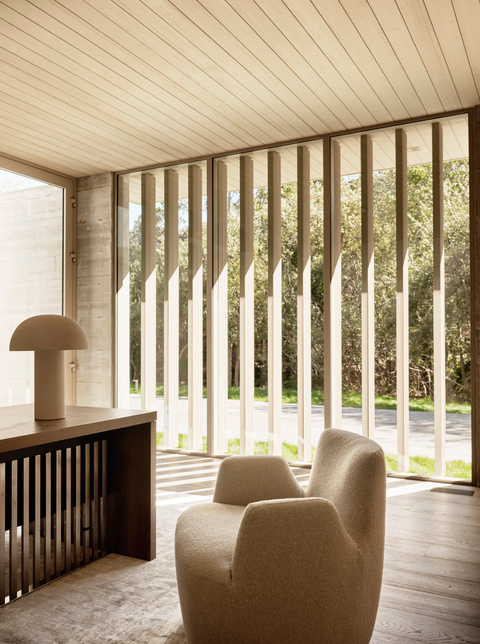 Cozy modern interior: armchair, desk with lamp, vertical blinds filtering sunlight, and exterior trees.