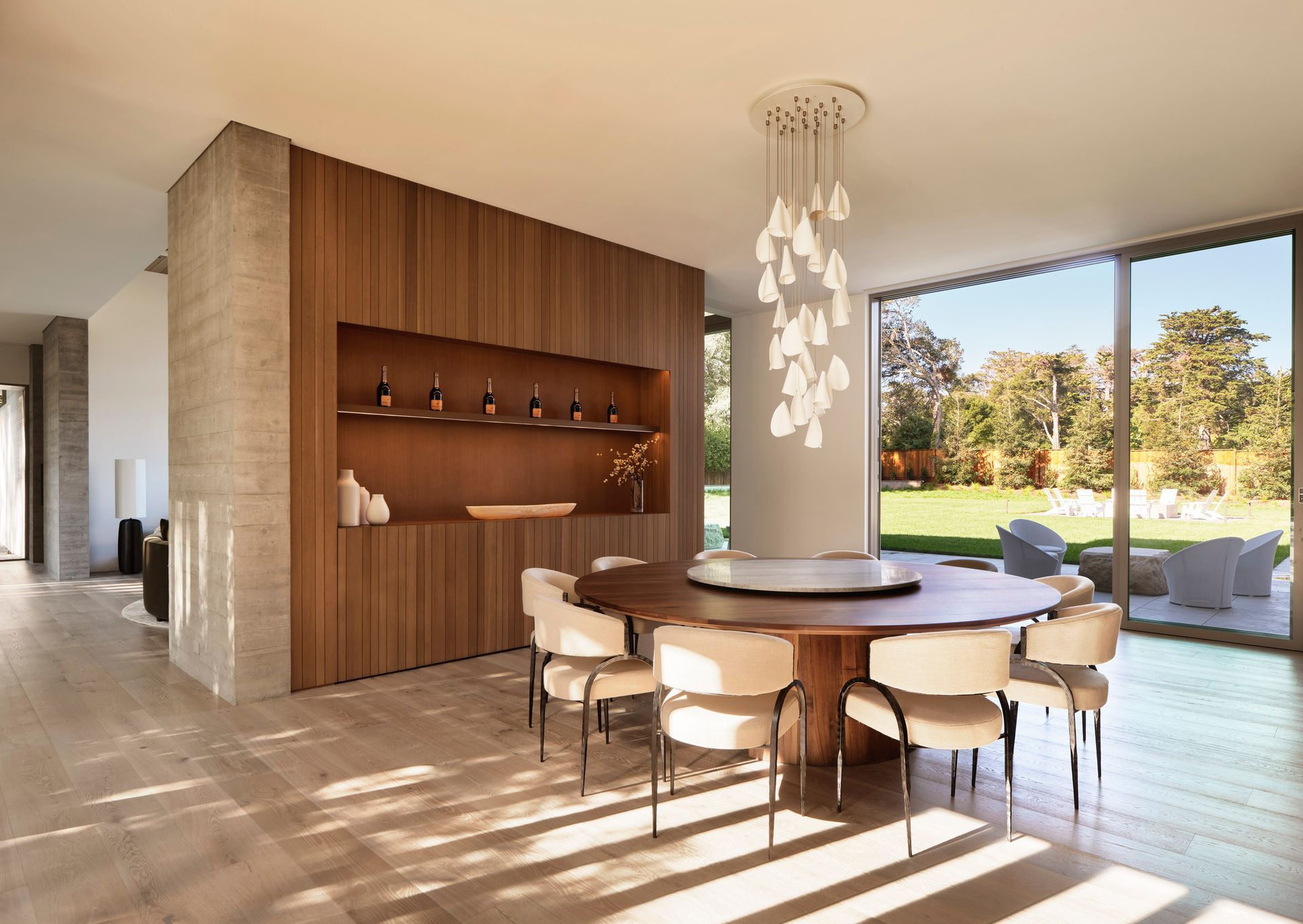 Modern dining room with a round wooden table, white chairs, and a chandelier, with large windows overlooking a yard.