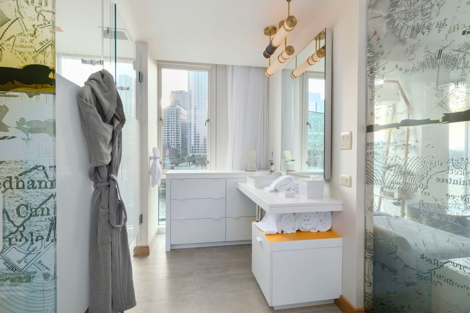 Bright bathroom with a gray robe hanging on the left; white sink and storage; city view through the window.