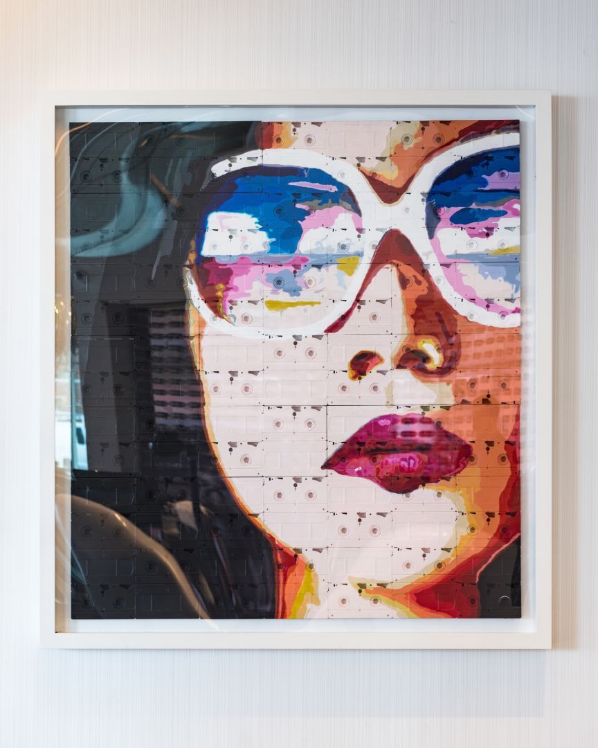 Portrait of a woman wearing sunglasses, the reflection in the glasses shows a colorful sky; framed art.