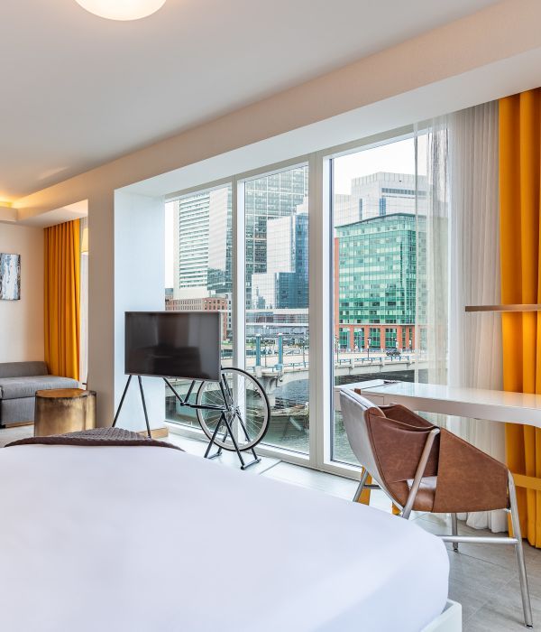 Modern hotel room with city view, large windows, white walls, and yellow curtains.