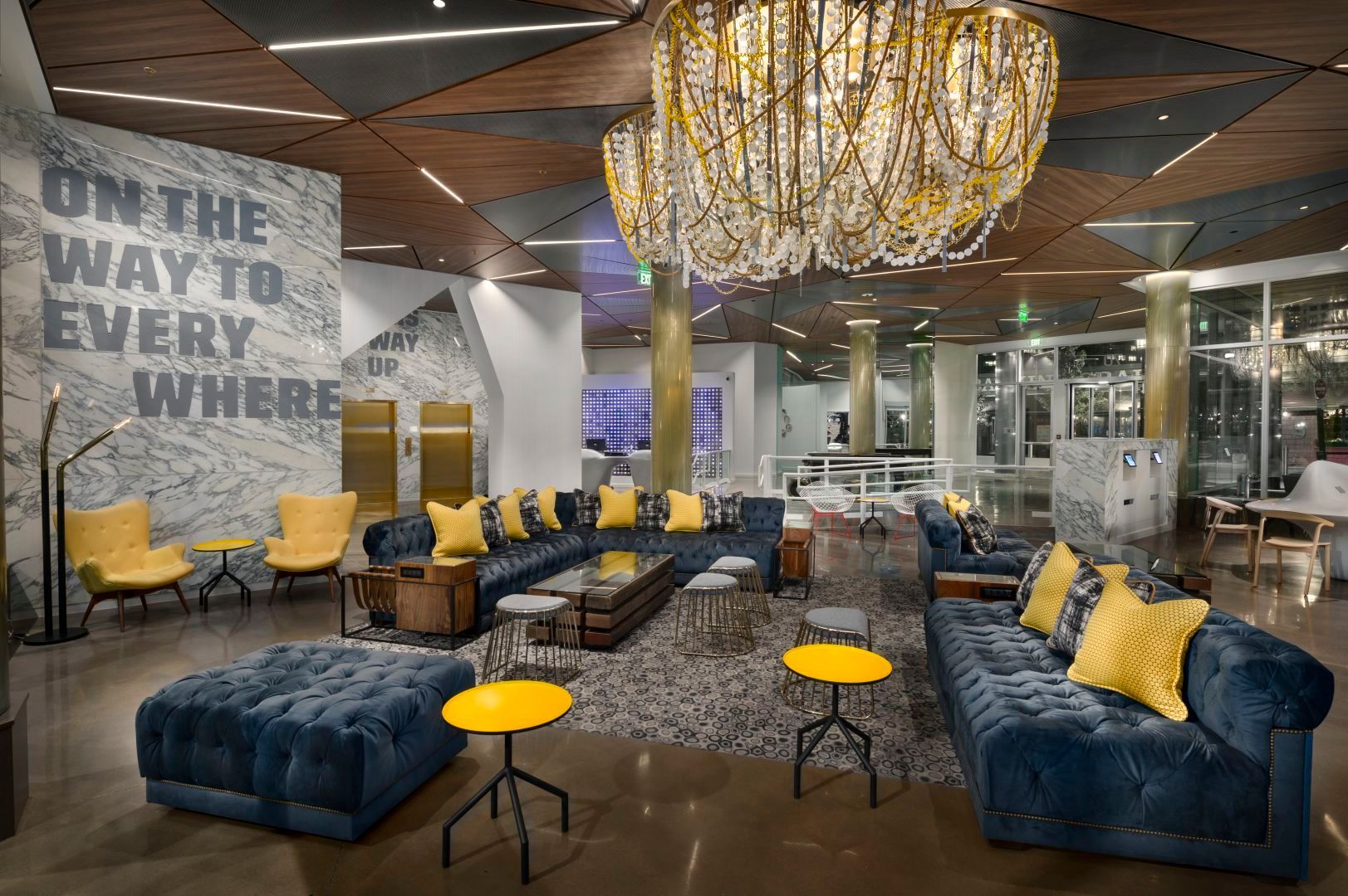 Lounge area with blue velvet seating, yellow accents, large chandelier, and wall text: 