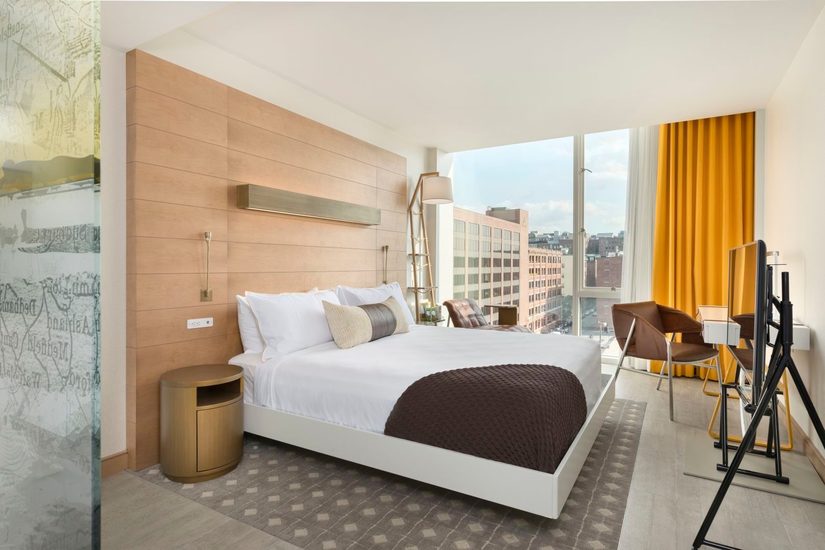 Bedroom with a large bed, brown accents, and a city view through a large window.