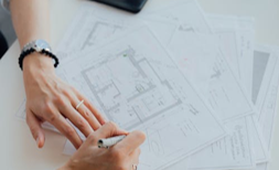Person's hands with watch and ring, drawing on floor plans with a pen at a white desk.