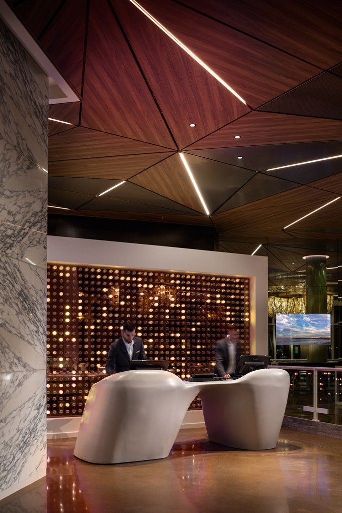 Modern hotel reception desk with two employees, brown wood ceiling with lights, and decorative wall.