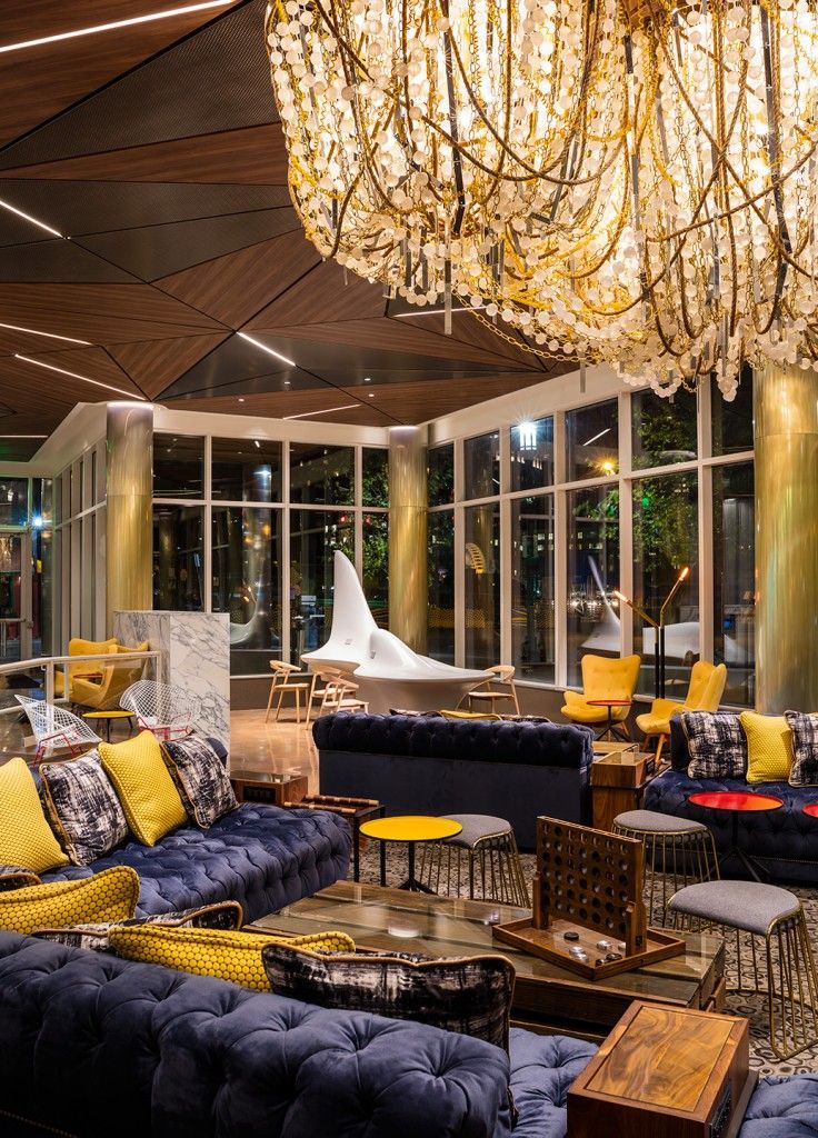 Lounge with blue sofas, yellow accents, and ornate chandelier. Large windows, modern decor.