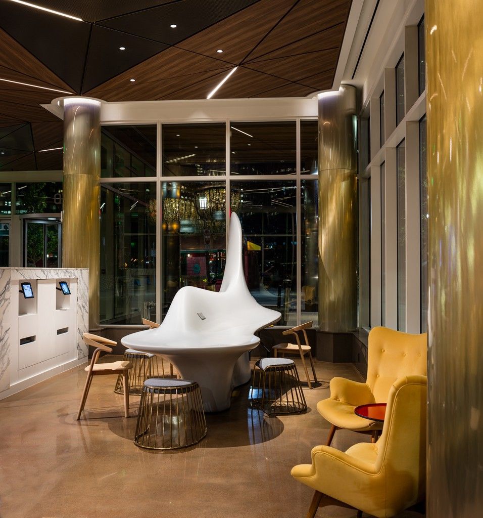 Lobby with sculpture, yellow armchairs, and gold pillars. Large windows offer a city view at night.