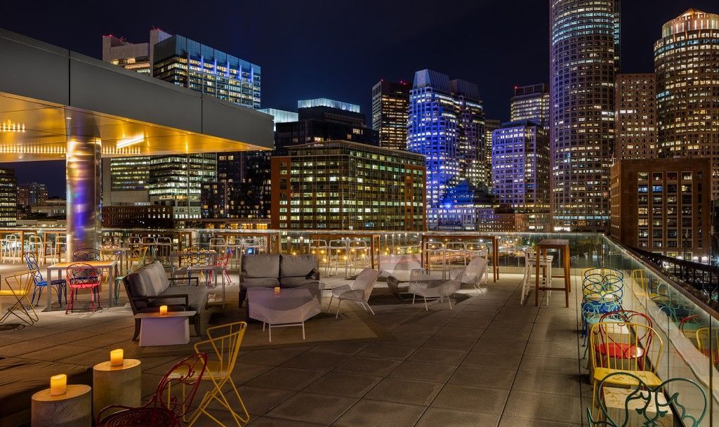 Rooftop bar at night with city skyline background. Tables, chairs, and lights are set up.