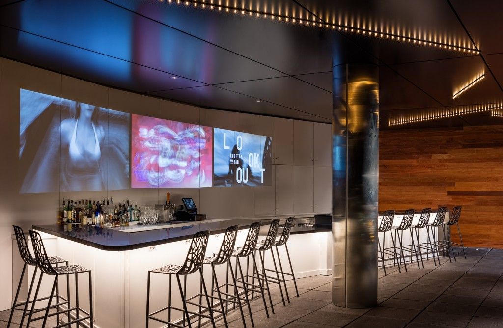Bar with seating, three screens, and a silver column. Lighting on the ceiling and a dark color scheme.