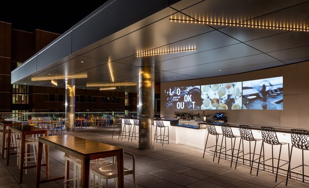 Outdoor bar with overhead lighting and projected images. Long wooden tables and bar seating.