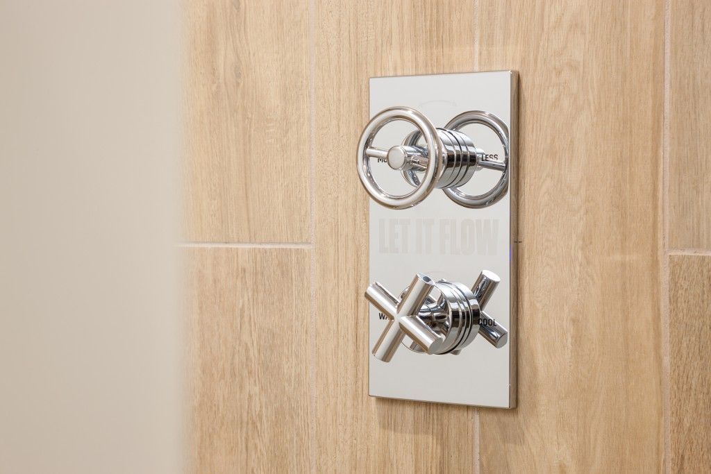 Chrome shower faucet with two wheel-style handles and one cross-style handle on a light wood tile wall.
