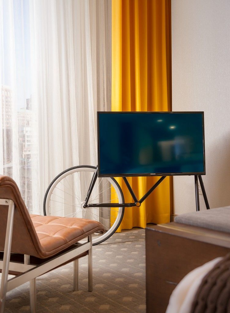 TV mounted on a bicycle frame stand in a room with a window and orange curtain.
