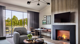 Cozy living room with fireplace, gray armchair, small dining table, and large window overlooking a balcony.