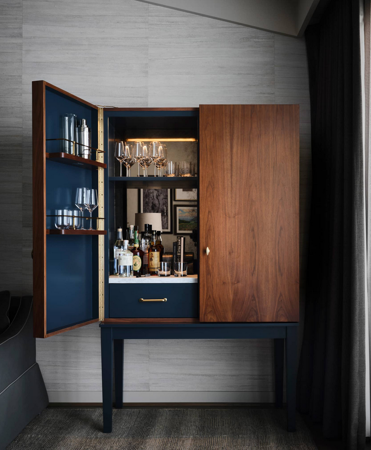 Wooden bar cabinet, one door open. Blue interior, shelves with glassware and bottles, with wood and gold accents.