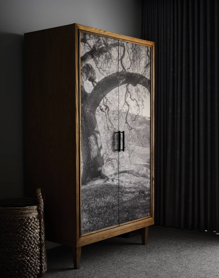 Wooden cabinet with a black and white tree image on the doors, in a room with a dark curtain and a woven basket.