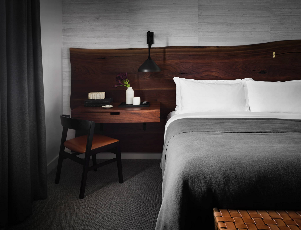 Bedroom with bed, desk, and chair. Dark wood headboard, gray blanket, black curtain, and neutral walls.