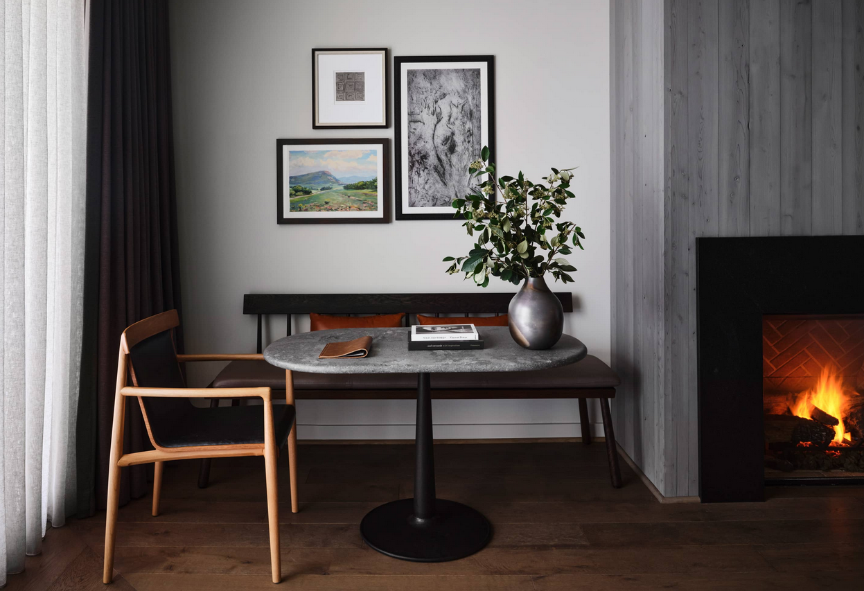 Cozy interior with a table, chair, bench, artwork, and fireplace. A vase with greenery sits on the table.