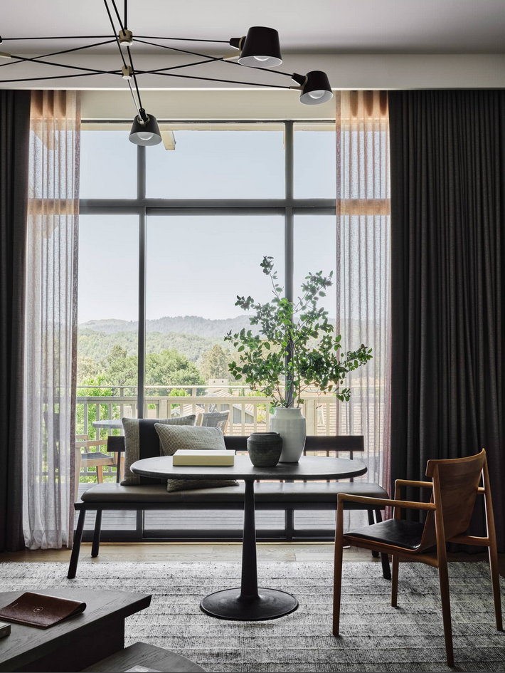 Dining area with a round table, chairs, and a bench in front of a window overlooking a green landscape.
