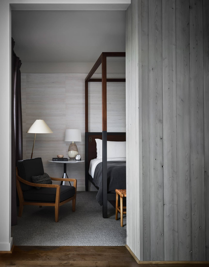 Bedroom interior with dark wooden bedframe, chair, and side table, seen through doorway.