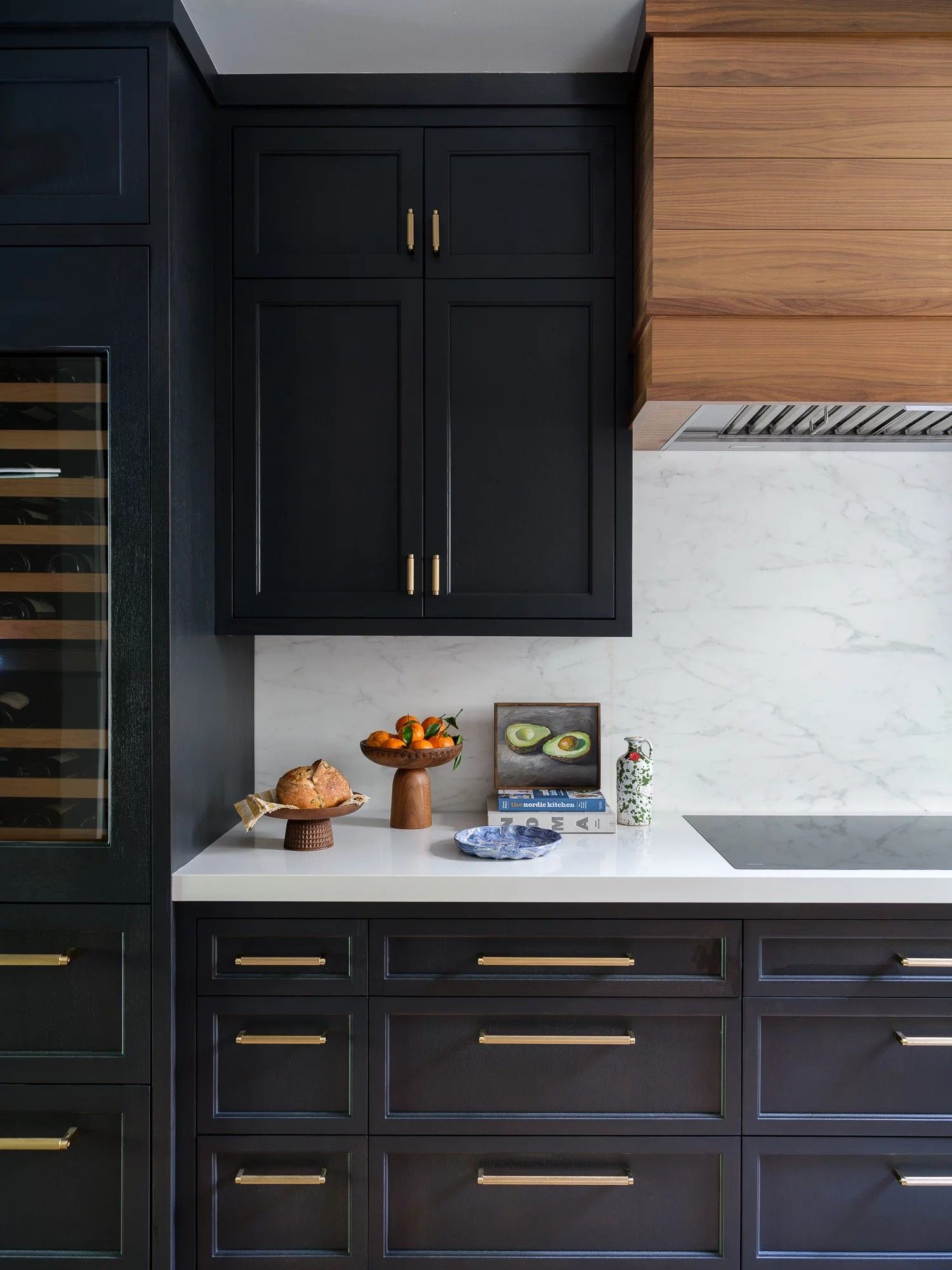 Dark kitchen with black cabinets, white countertops, wood range hood, and gold hardware.