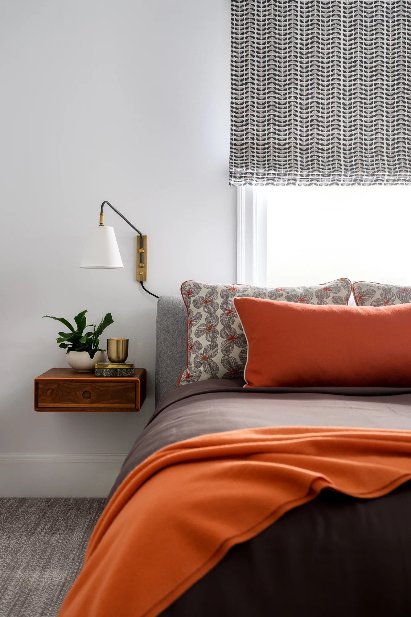 Bedroom with bed, floating nightstand, wall-mounted lamp, patterned Roman shade, orange accents.