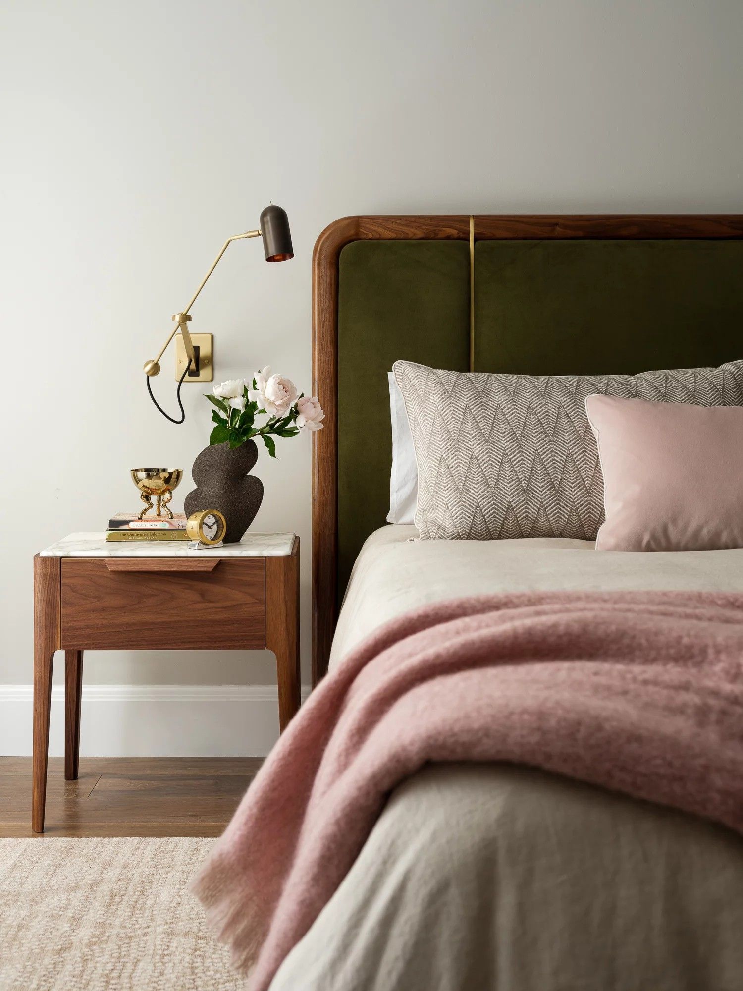 Bedroom with olive green headboard, wooden nightstand, pink blanket, and floral arrangement.