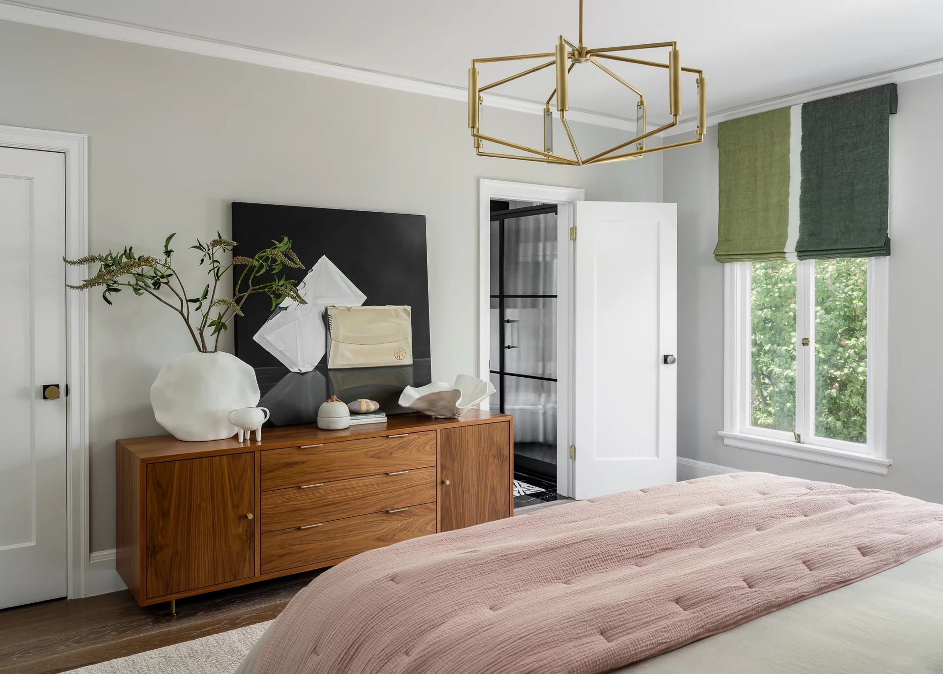 Bedroom with wooden dresser, artwork, and window with green and tan shades; pink blanket on bed.