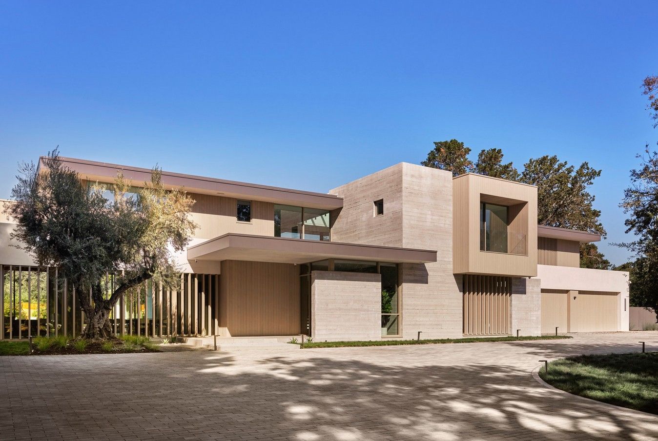 Modern house with light-colored brick exterior, wooden accents, and a long driveway under a blue sky.