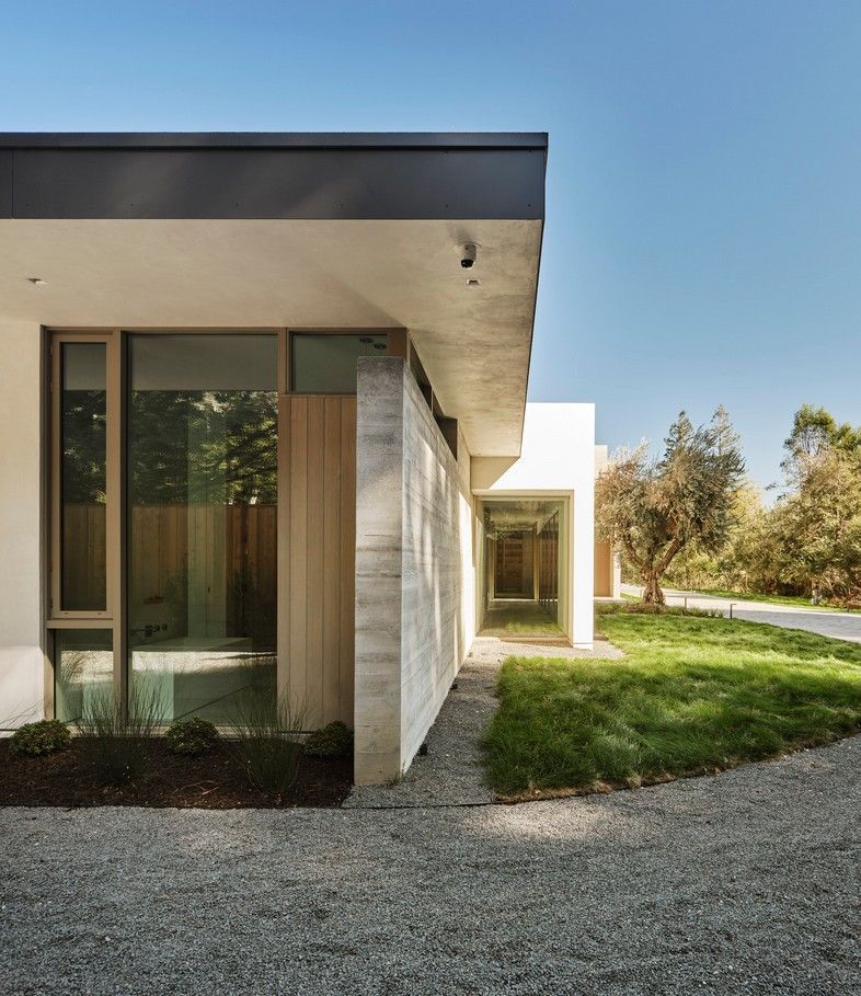 Modern house exterior with concrete and glass walls, gravel path, and greenery under a blue sky.