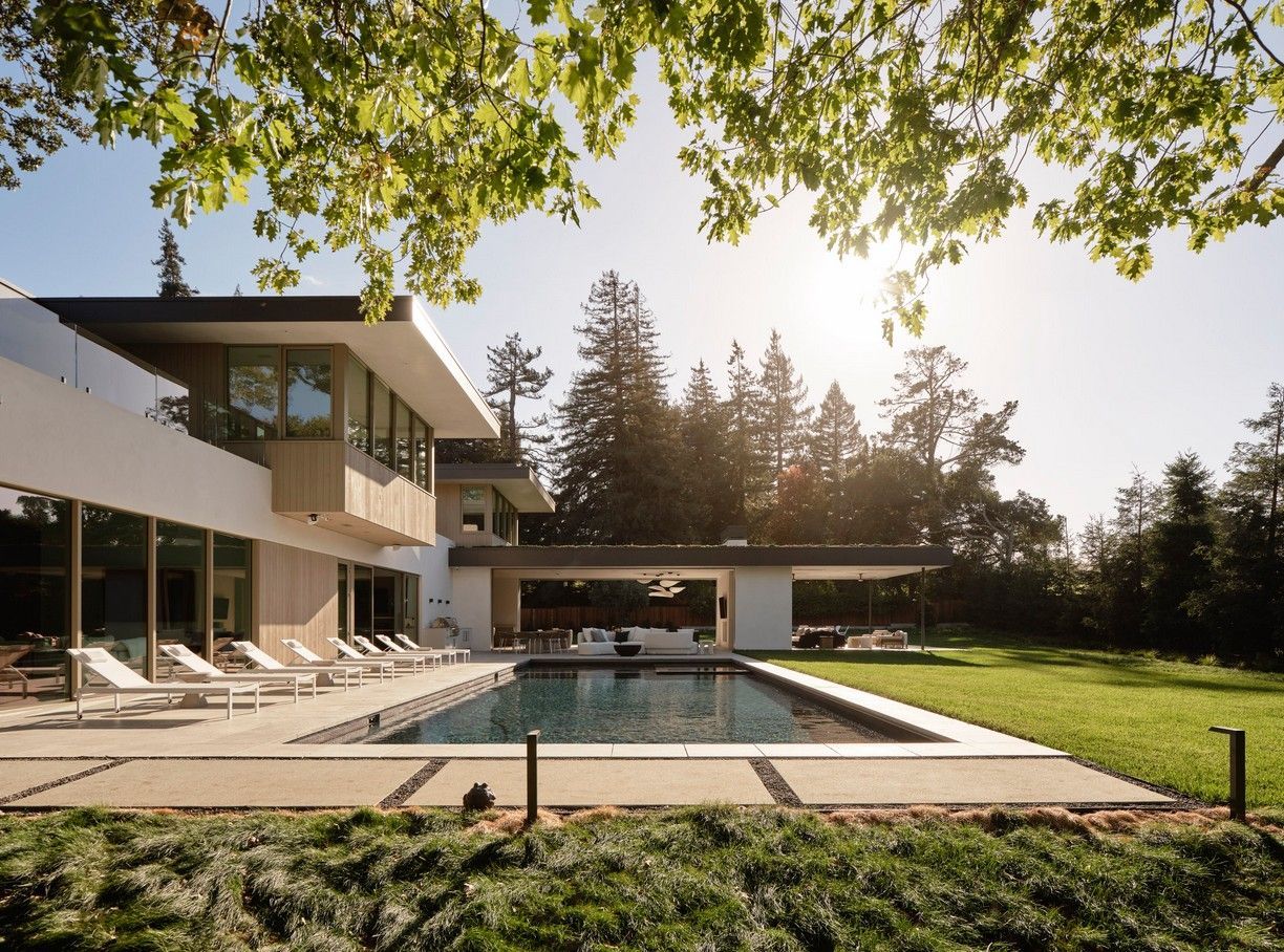 Modern house with a pool and lounge chairs on a sunny day. Green lawn and trees.