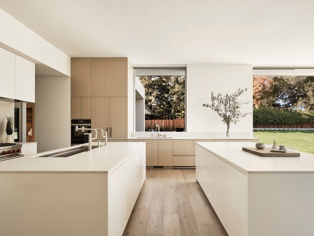 Modern kitchen with two white islands, light wood floor, and large windows overlooking a yard.