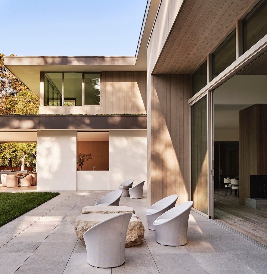 Modern home exterior with outdoor seating on a stone patio. Features include large windows and minimalist design.