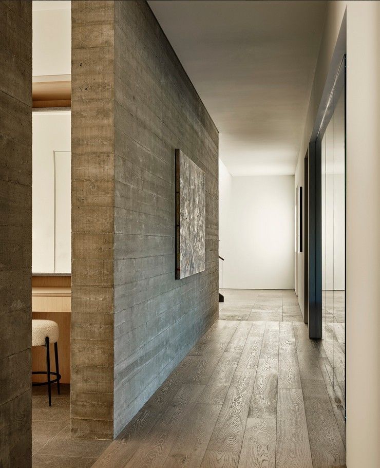 Long hallway with textured grey wall and light wood flooring; art piece and open doorway visible.