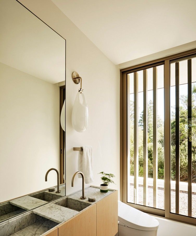 Modern bathroom with a large mirror, dual sinks, and vertical blinds overlooking greenery.