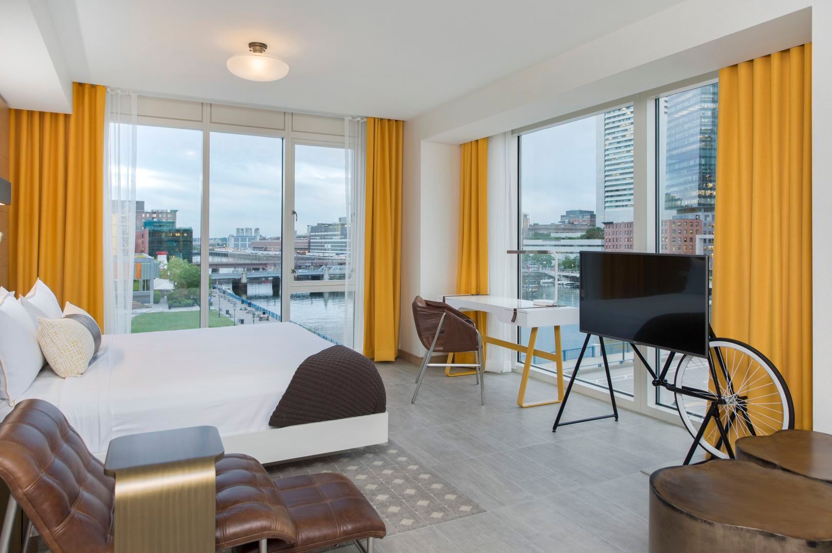 Hotel room with bed, large windows, yellow curtains, city view, desk, TV.