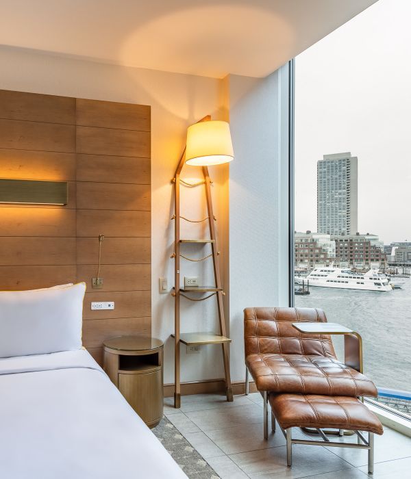 Hotel room with a bed, a leather chair by a window, and a unique lamp with shelving. View of the water and tall building.