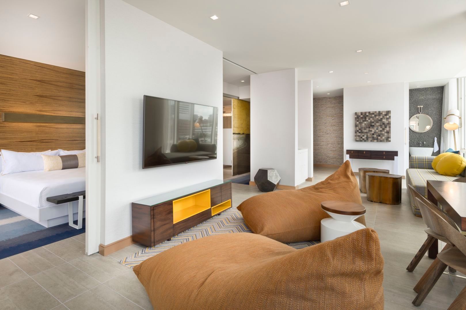 Modern hotel suite with living area and bedroom visible; brown beanbag chairs, wood accents, and a mounted TV.