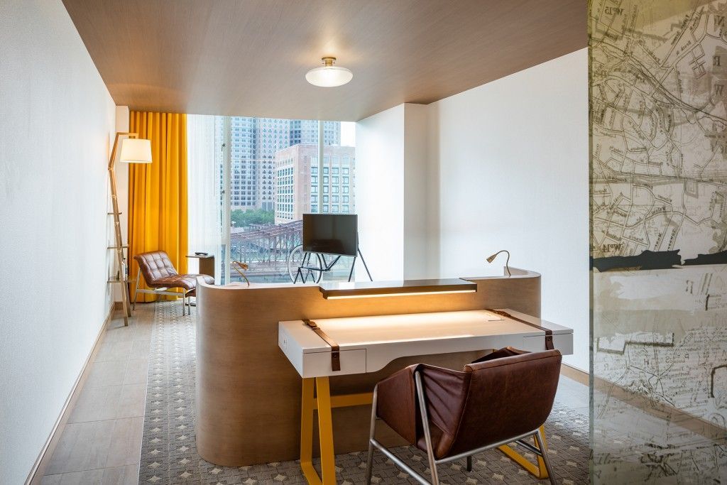 Hotel lobby with a curved reception desk, yellow chair, and floor-to-ceiling windows overlooking a city.