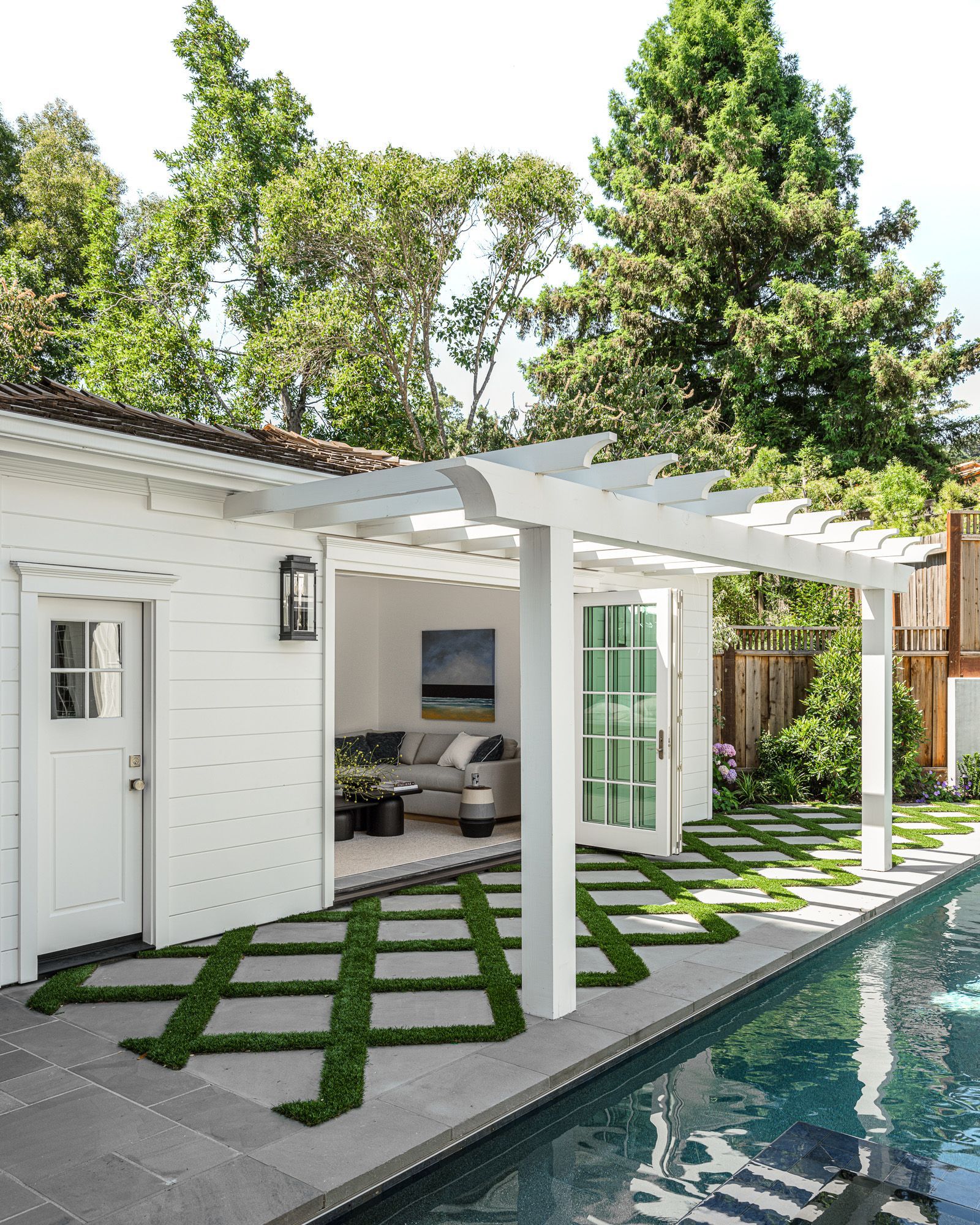 White pool house with pergola, door and windows, next to a pool. Green grass and trees in background.