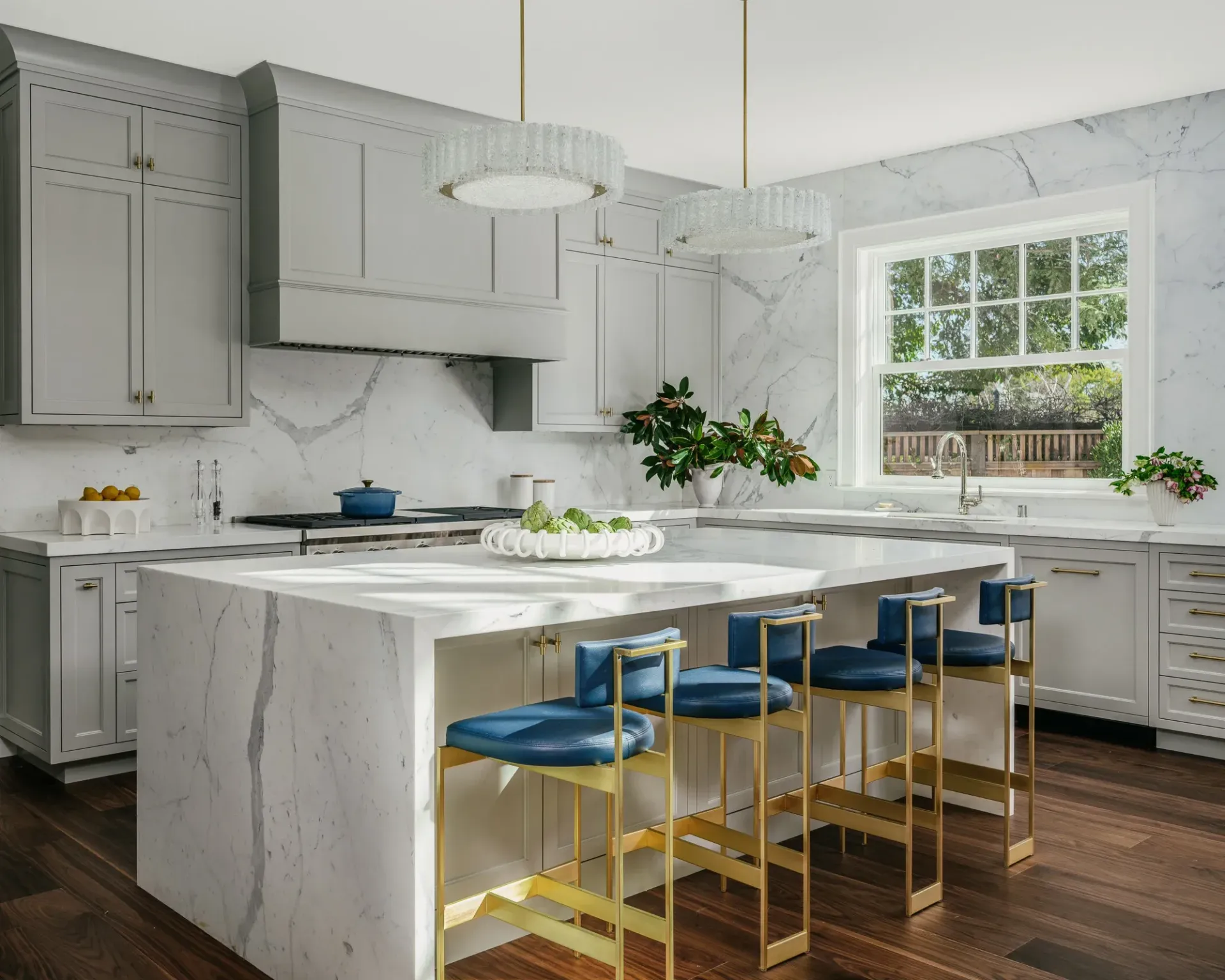 Modern kitchen with marble countertops, gray cabinets, blue stools, and natural light.