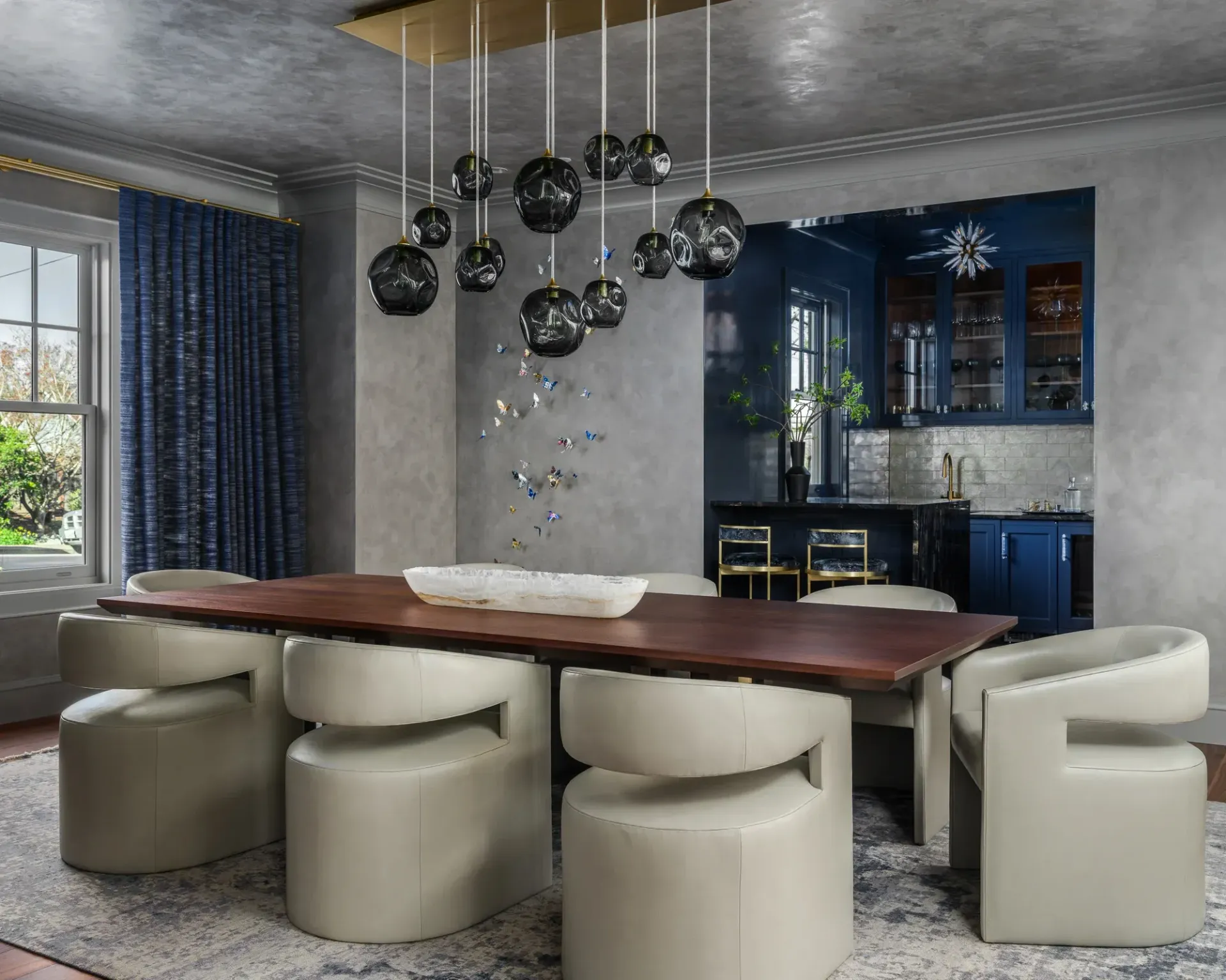 Elegant dining room with a long wooden table, cream chairs, and a dark blue kitchen in the background.