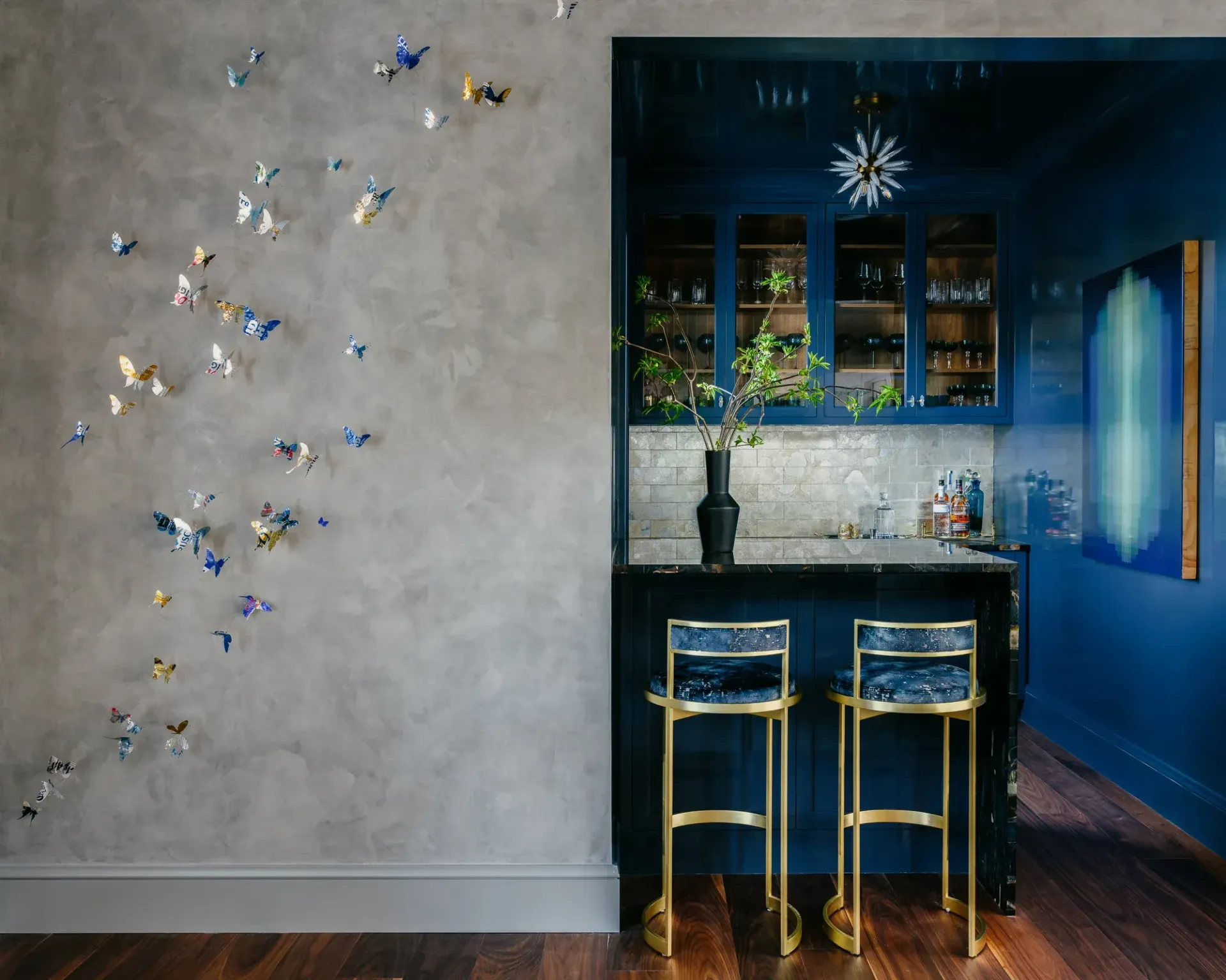 Gray wall with butterfly art beside a dark blue bar. Gold stools and a painting add color.