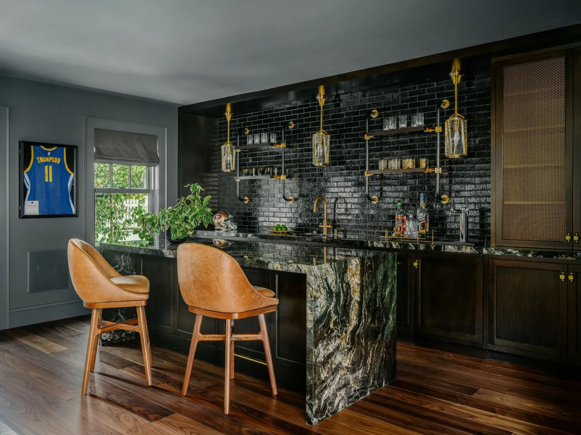 Luxury bar with green marble countertop, black backsplash, gold accents, two leather stools, and a framed jersey.
