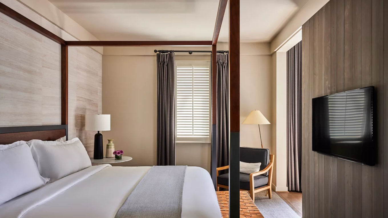 Bedroom with canopy bed, neutral tones, wood accent wall, armchair, and window with blinds.